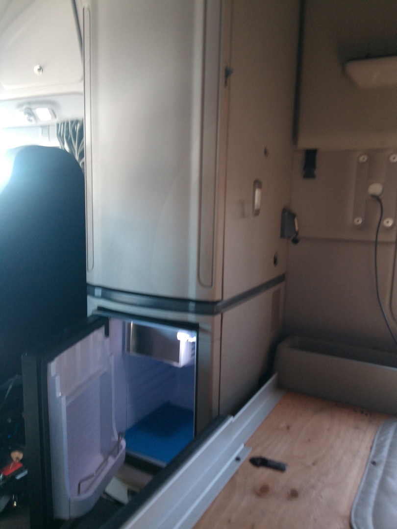 Truck cab interior showing open fridge/freezer with blue interior and storage area; vertical storage cabinet with a freezer at lower left.