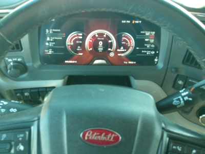 Peterbilt dashboard view from steering wheel showing digital gauges in red; Peterbilt emblem on steering wheel; faint left and right stalks; center digital display.