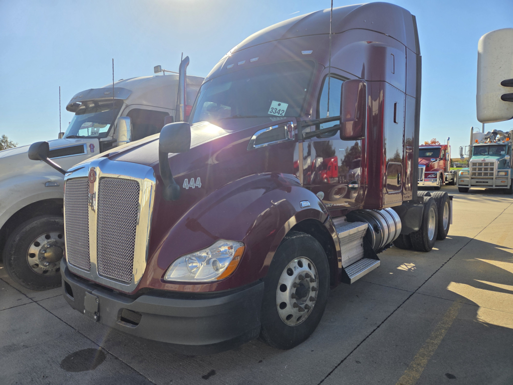 Kenworth T680 tractor-trailer in burgundy showing aerodynamic sleeper cab and dual rear axles; front grille with Kenworth badge; side fuel tanks; daylight; other trucks in background.