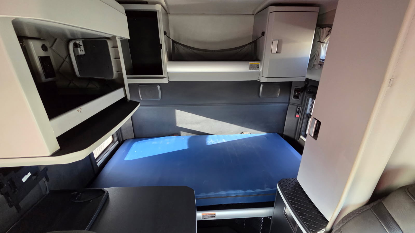 Kenworth T680 sleeper cab interior showing bunk area with blue mattress and surrounding cabinets and storage compartments.