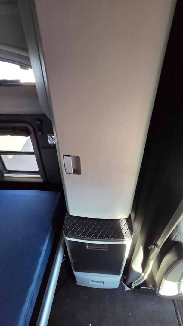 Truck interior side panel and step area of a 2022 Kenworth T680, showing a small storage drawer and metal diamond-plate step near a beige vertical panel.