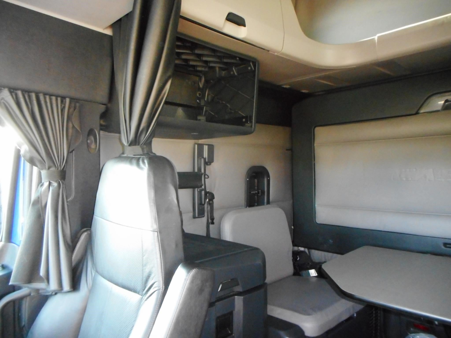 Truck cab interior of a 2022 Freightliner Cascadia 126 showing the sleeper area with a small table, curved window, and overhead storage.