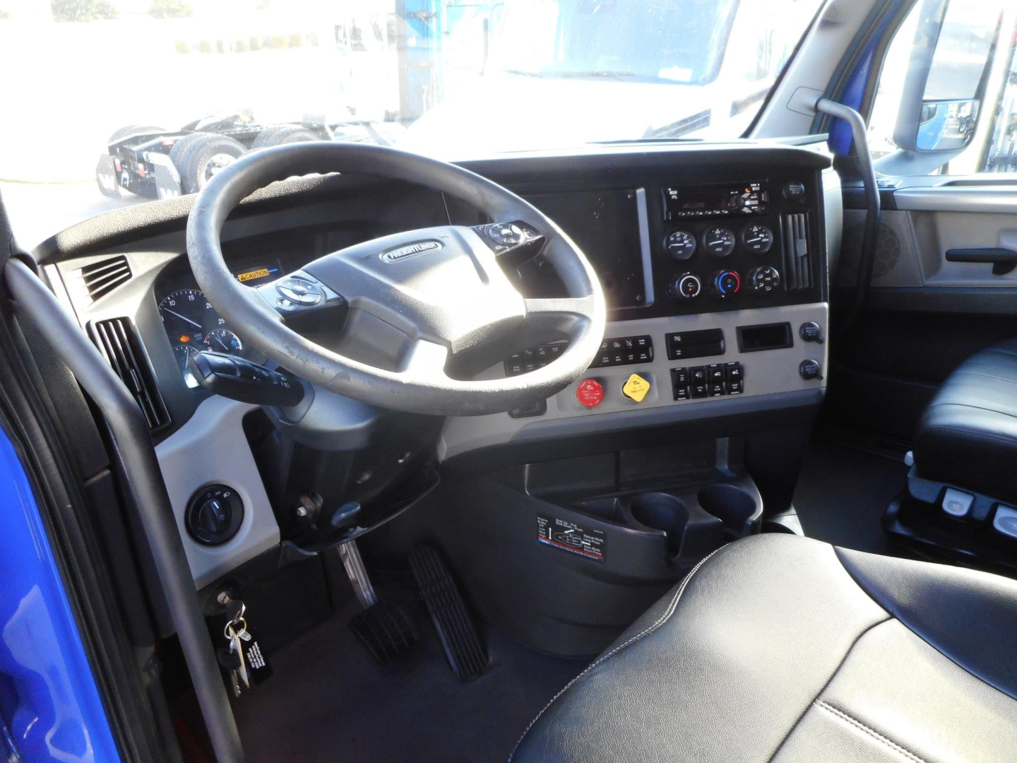 Truck cab interior showing steering wheel, dashboard gauges, touch-screen display, and center console of a Freightliner Cascadia. Visible pedals and ignition area.