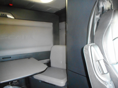 Truck cab interior of a 2022 Freightliner Cascadia 126 showing sleeper area with table and seat, light gray upholstery, right-side view of passenger seat, overhead storage panel.