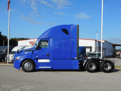 Freightliner Cascadia 126 semi truck in royal blue, side view showing sleeper cab and triple rear axles, dealership lot in background.