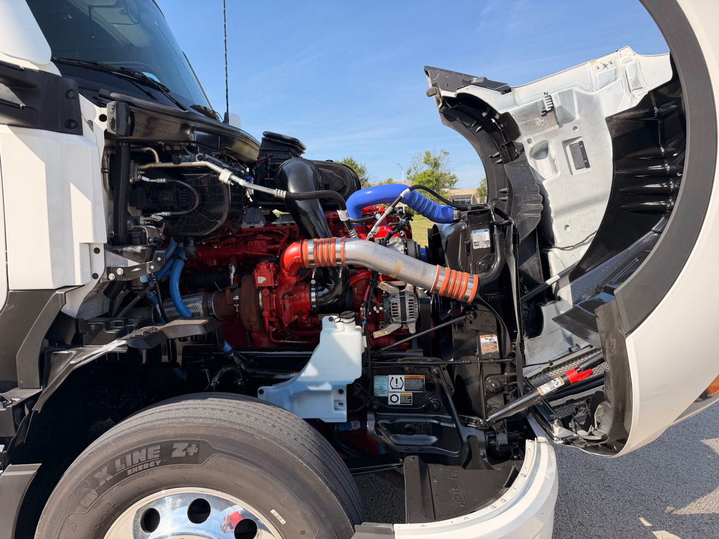 Kenworth T680 engine bay with red engine, blue hoses, and exposed chassis; front left wheel visible; cab open showing engine components.