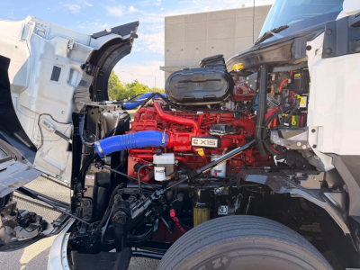 Kenworth T680 engine bay with red Cummins engine and exposed components, blue intercooler hose, front wheel visible, cab-off approach.