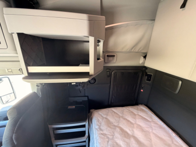 Kenworth T680 sleeper cab interior showing bunk area with overhead storage and small cabinet; cockpit partially visible with light gray interior panels.