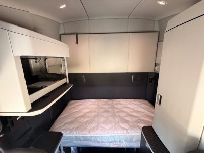 Kenworth T680 sleeper cab interior showing bed with mattress, cabinets, and side storage panels in a gray-white cabin.