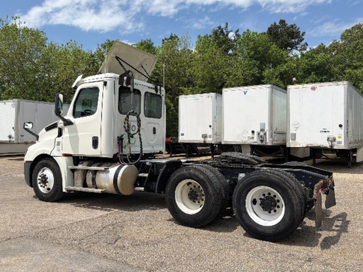 Truck cab and chassis of a 2019 Freightliner Cascadia126 in a parking lot, white cab, exposed engine hood raised, multiple tandem rear axles, with cargo trailers in background.