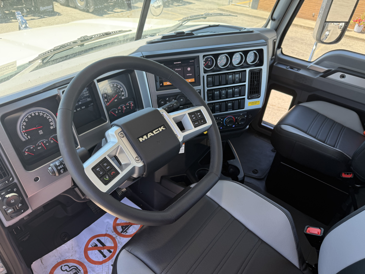 Dashboard and steering wheel of a Mack PI84T truck cab showing steering wheel with 'MACK' logo, gauge cluster, control panels, and center stack with vents and buttons.