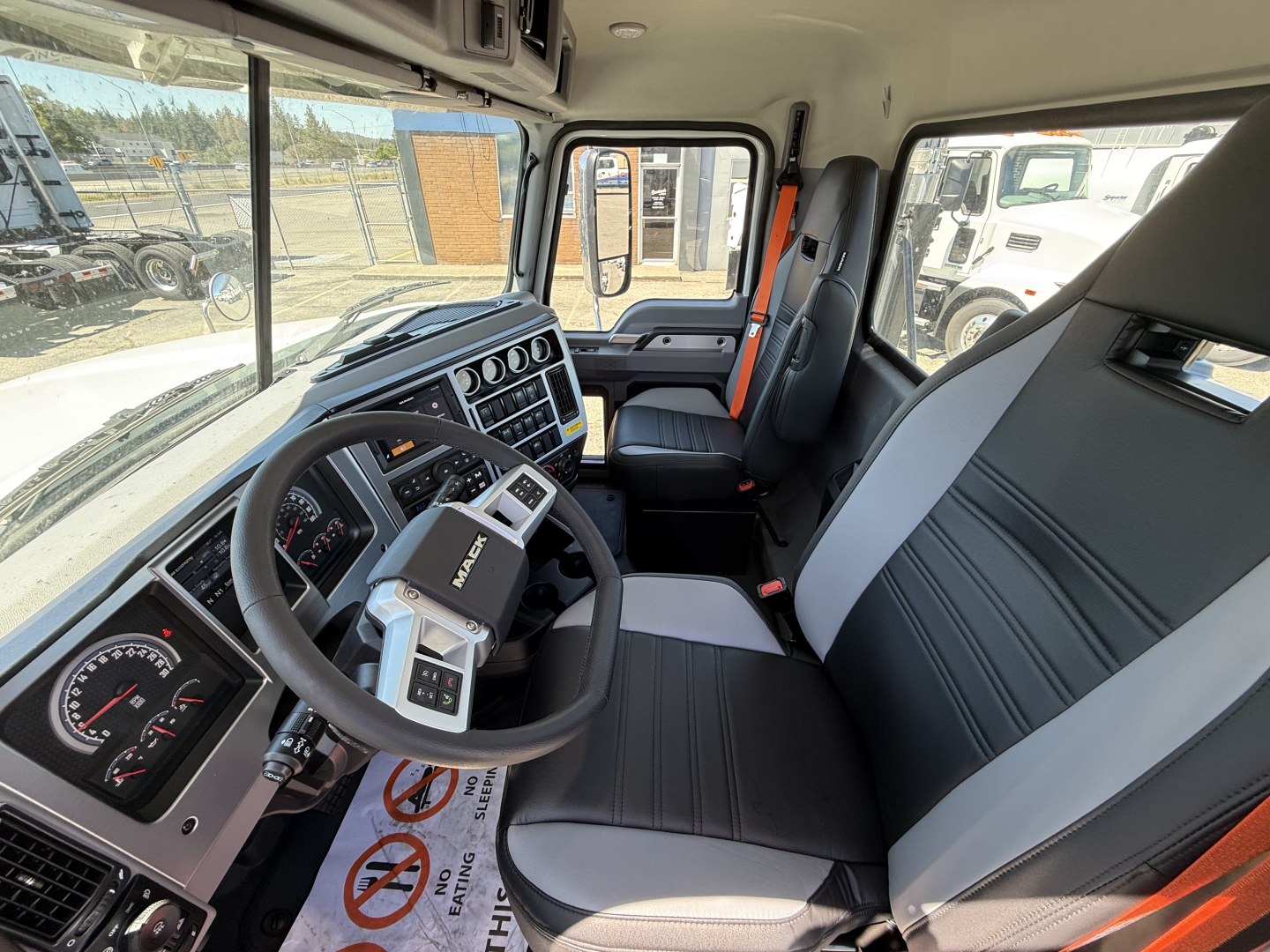 Truck cab interior showing steering wheel, dashboard gauges, center console, and two-tone black and gray seats with orange seat belts. Visible text on controls: Mack steering wheel badge.