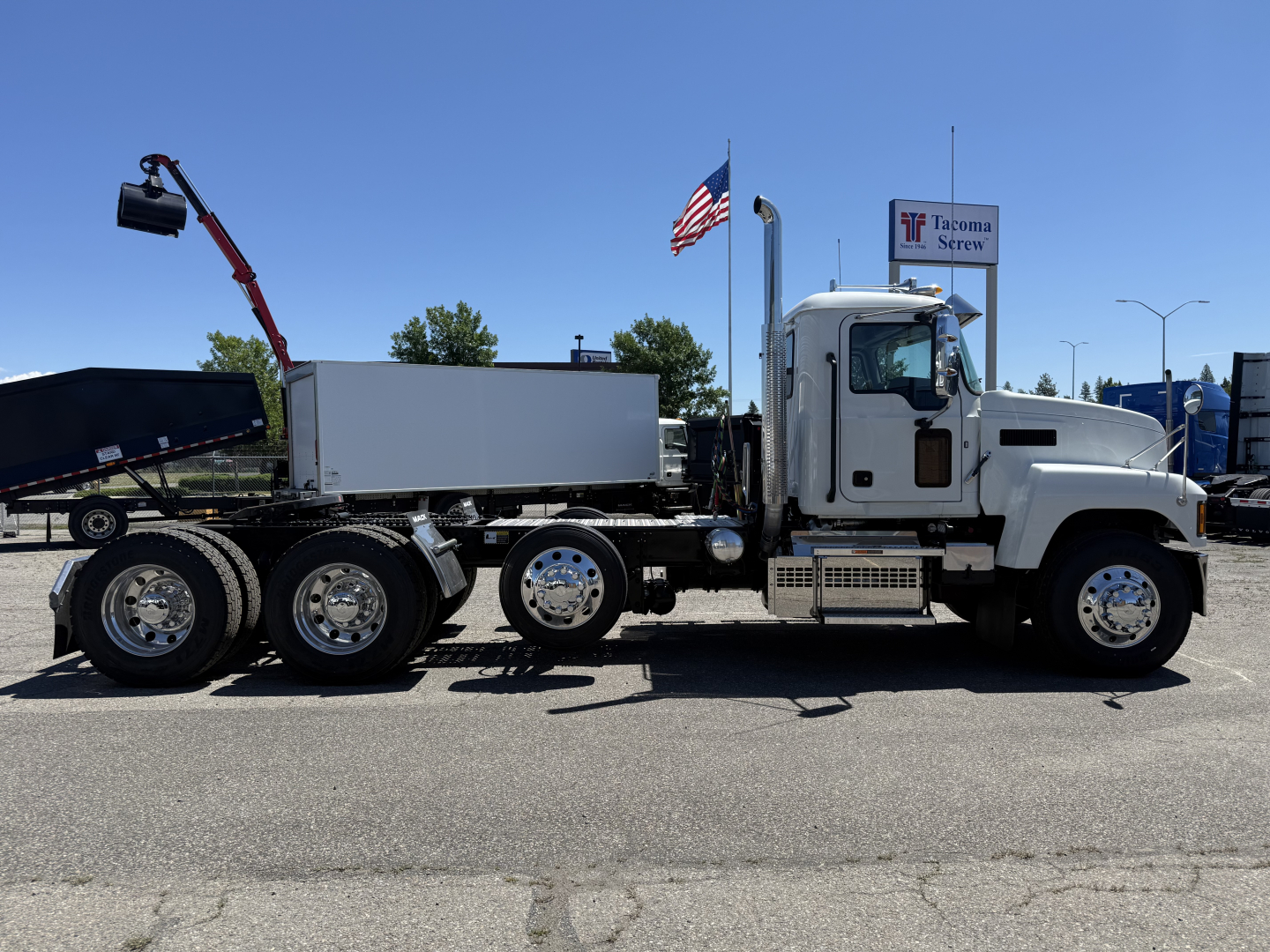 A white Mack PI84T semi-truck in profile view with a flatbed/dump behind, dual rear axles, chrome wheels, and a small cab; an American flag and Tacoma Screw dealership sign visible.