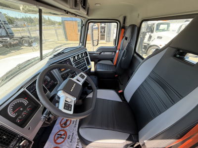 Truck cab interior showing steering wheel, dashboard gauges, center console, and two-tone black and gray seats with orange seat belts. Visible text on controls: Mack steering wheel badge.