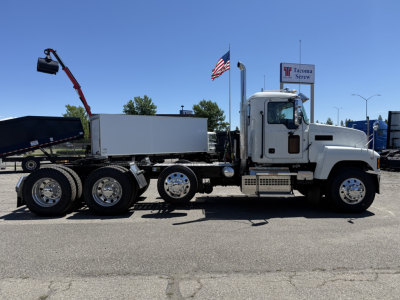 A white Mack PI84T semi-truck in profile view with a flatbed/dump behind, dual rear axles, chrome wheels, and a small cab; an American flag and Tacoma Screw dealership sign visible.