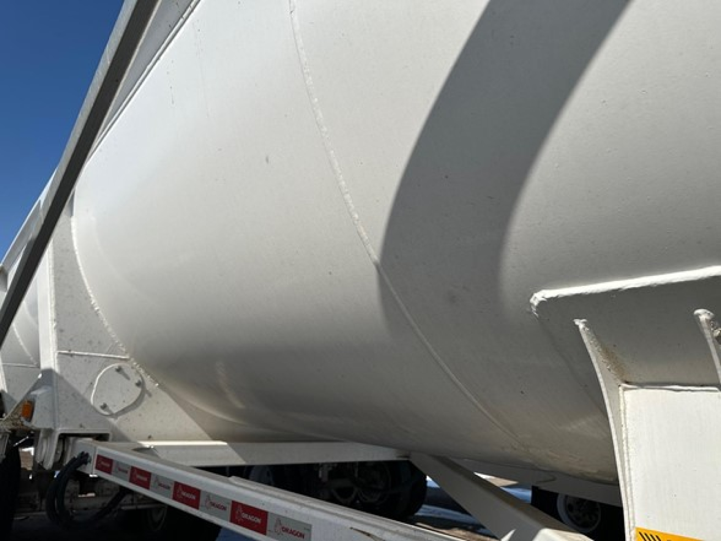 Dump trailer side angle showing curved tanker-like aluminum body and white frame; visible wheel and support structure; no text readable on the surface.