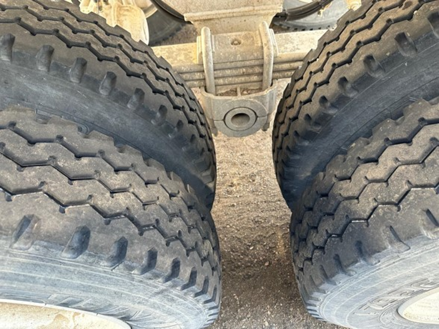 Trailer axle close-up showing dual rear tires with heavy tread, leaf spring suspension, and universal joint housing between wheels.