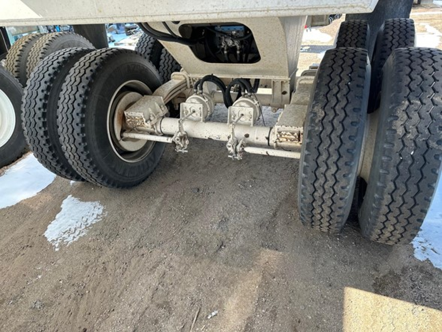 Trailer undercarriage showing dual axle suspension and leaf spring assemblies with tires, disk brakes, and central differential housing; metallic components with dirt and road grime.