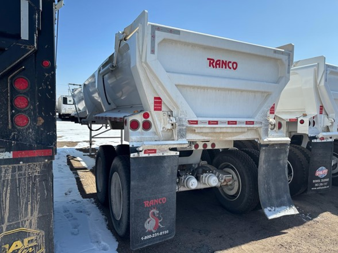 Dump trailer showing RANCO ED26-34 logo on white body with dual rear wheels and tailgate, parked among other trailers.