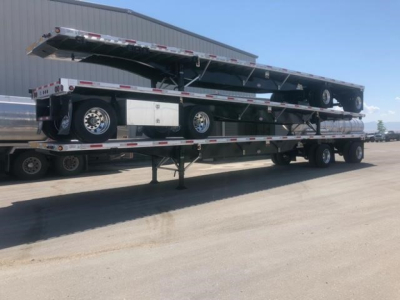 Utility 4000AE flatbed trailer on a multi-axle carrier with folded deck raised, showing tires, hydraulic components, and metal storage box.