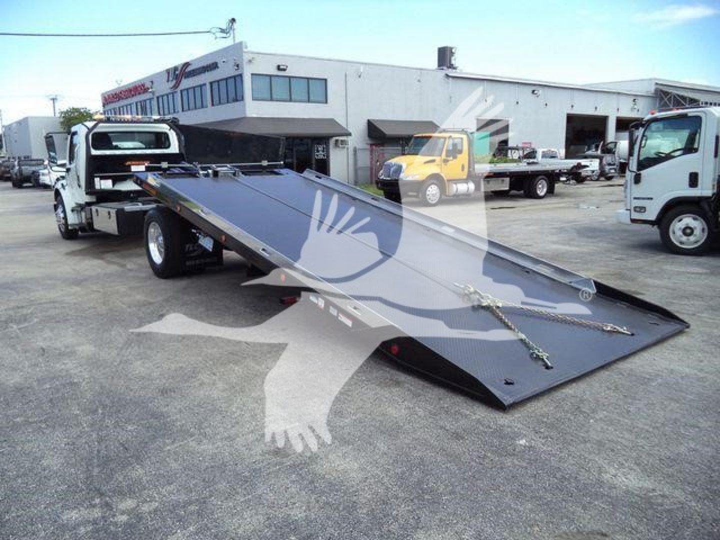 Flatbed tow truck with a lowered steel ramp in a dealership lot.