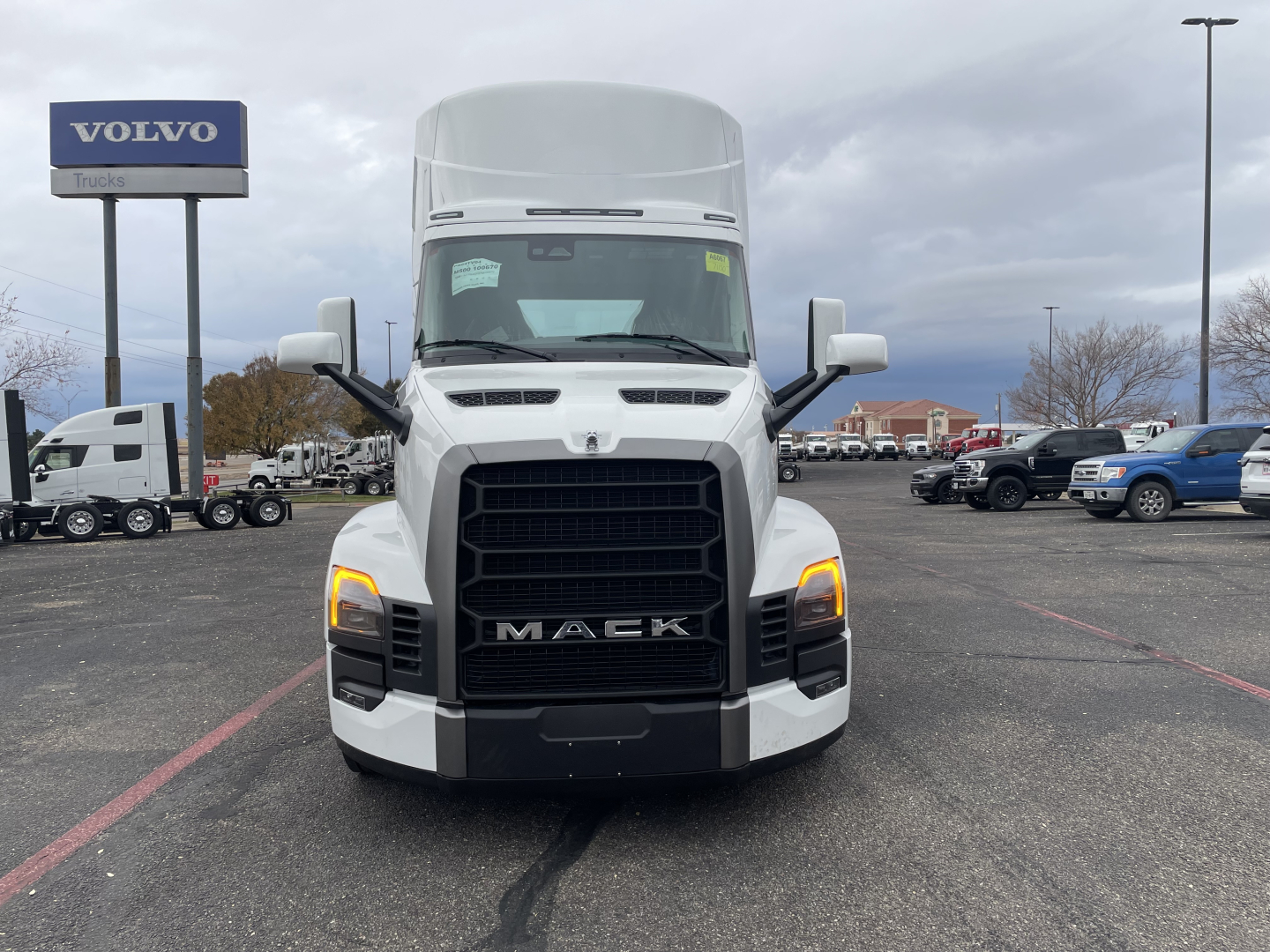Front view of a white 2026 Mack PR64T conventional daycab truck in a dealership lot with black grille and amber turn signals.