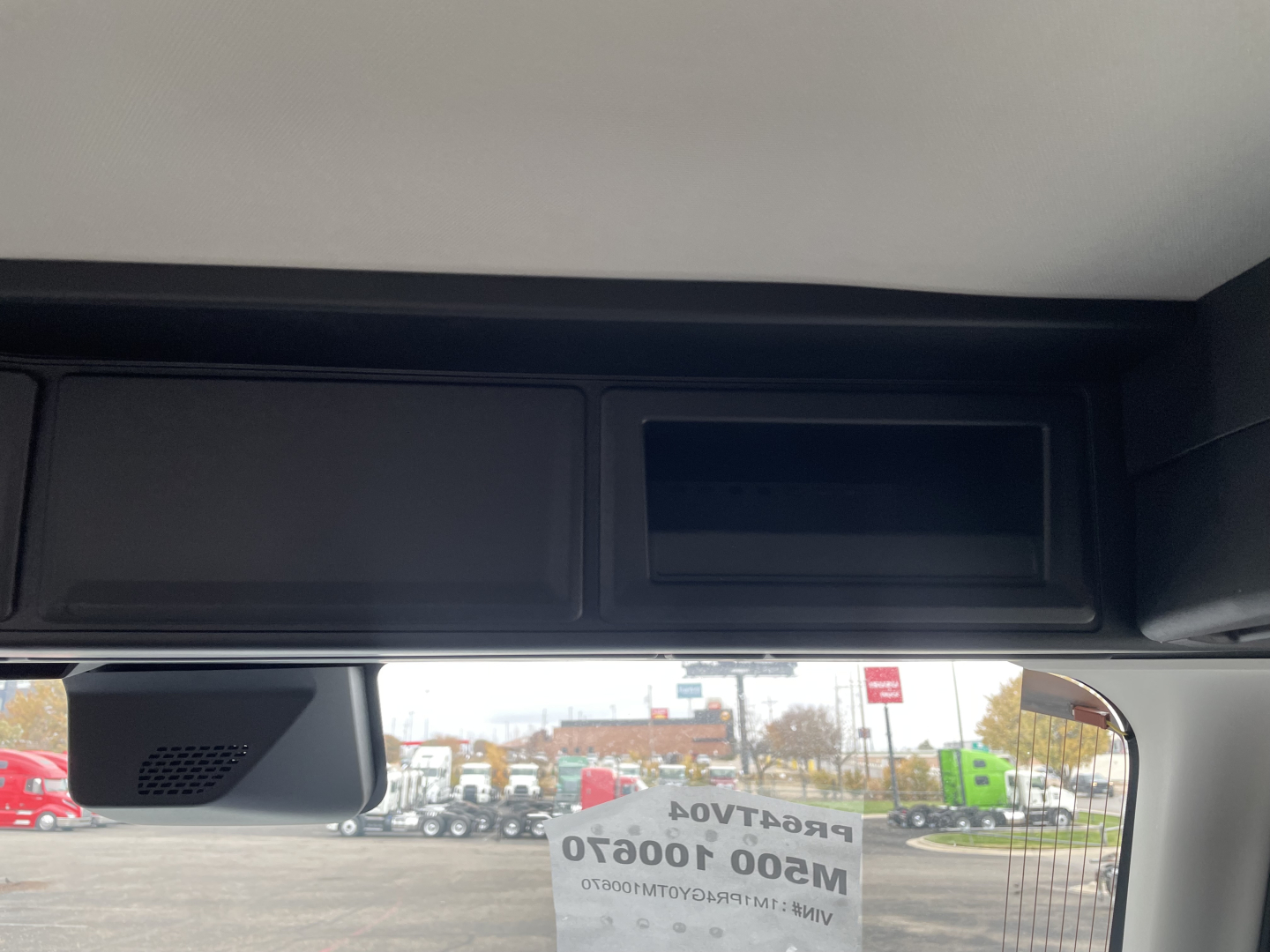 Truck cab interior showing overhead storage compartments and a forward-facing mirror; windshield with reflection of equipment lot.
