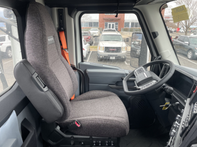 Truck cab interior with steering wheel and pilot seat of a 2026 Mack PR64T, grey fabric seat, dashboard, and gear controls visible.