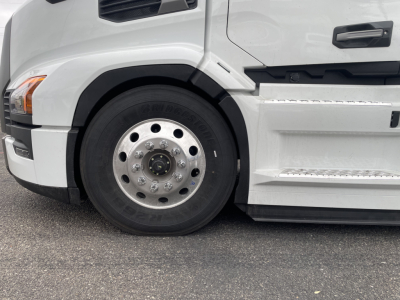 Truck wheel and front fender of a white Mack PR64T conventional daycab showing aluminum wheel with multiple lug nuts and tire details.