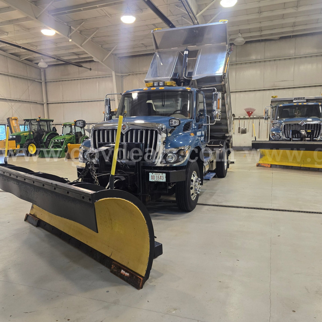 Snow plow-equipped 2013 International 7400 truck in a showroom with a large yellow and black plow blade attached in the foreground.