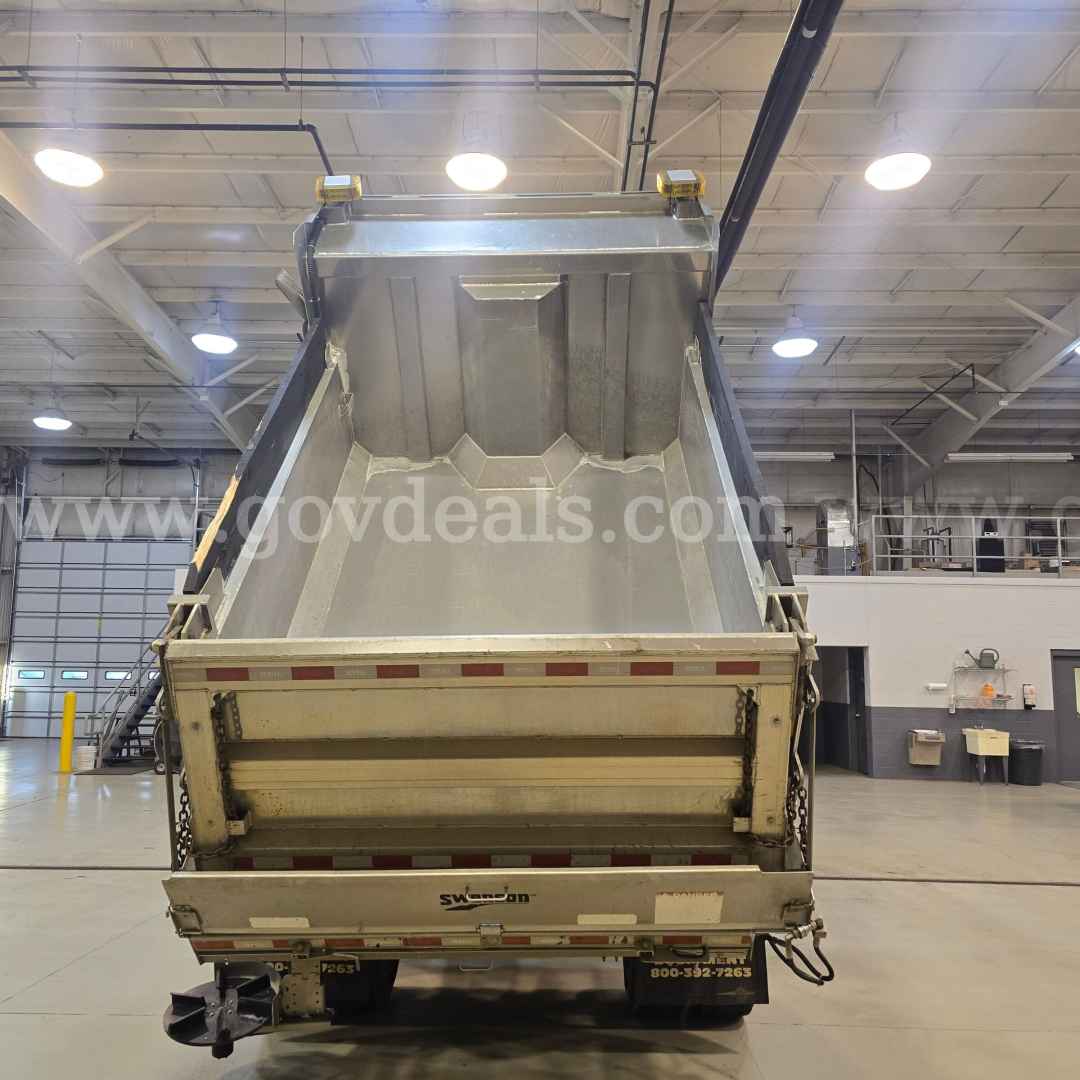 Dump truck bed with rear tailgate and plow equipment in a showroom, International 7400 model, tail lights and reflective tape visible.