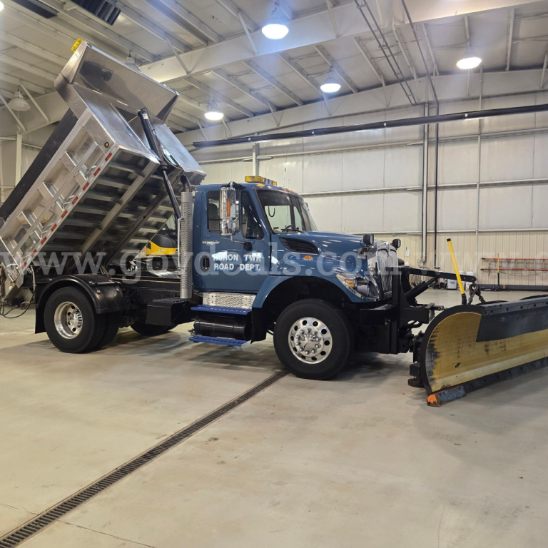 2013 International 7400 plow truck in a showroom with a front plow and a dump bed raised, blue cab, DOT markings on door, indoor lighting.