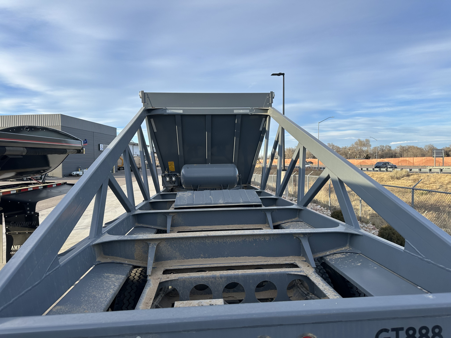 Dump trailer frame from rear with open center, wheel wells and hitch area. Steel beams, triangular supports, and a hopper with a cylindrical component visible.