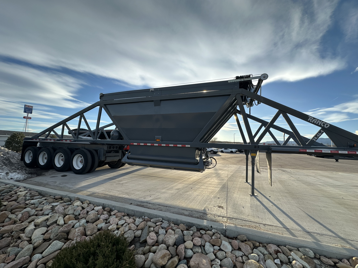 A dump trailer with a large gray hopper and multiple axles, shown in side view. No readable text visible on the trailer itself.