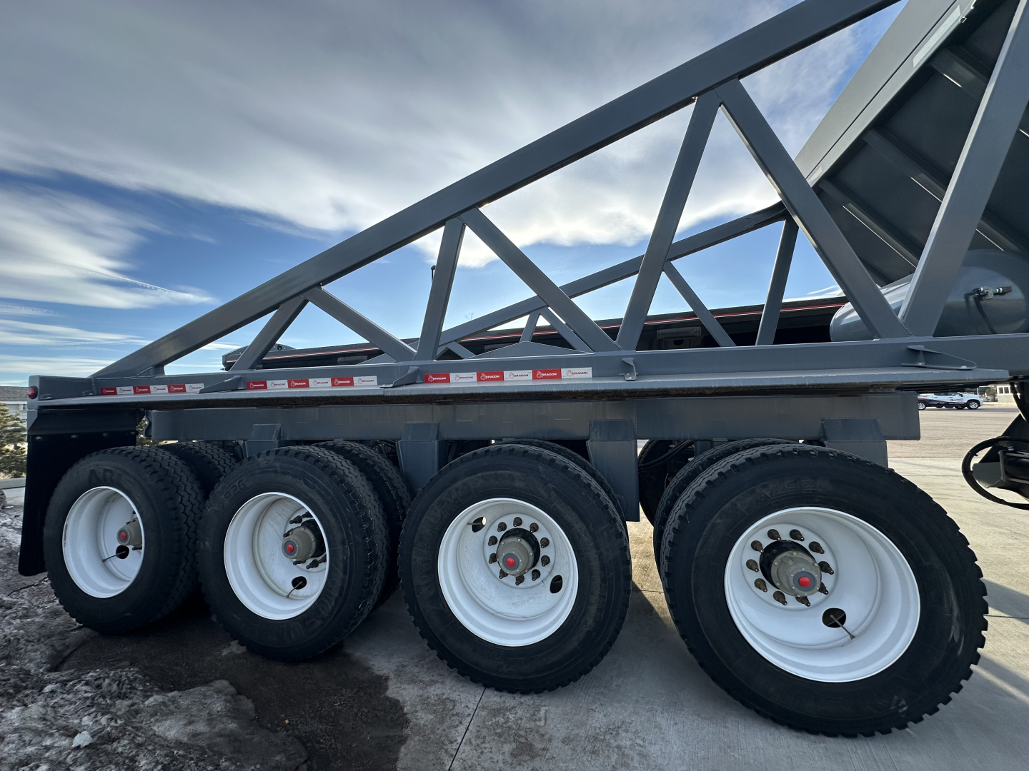 Trailer side view showing 6-wheel tandem axle with white wheels and black tires; steel frame with triangular lattice beams and reflective tape along the side.