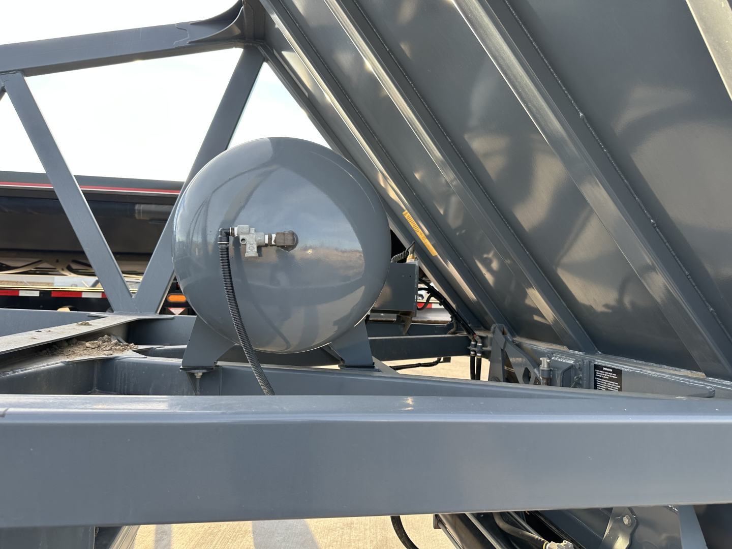 Dump trailer undercarriage showing a circular propane or air tank mounted on a metal frame with a raised bed in the background.