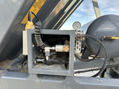 Dump trailer control panel with hydraulic hose, gauge, and wiring inside a metal housing; yellow warning label; hose connections and cylindrical components visible.