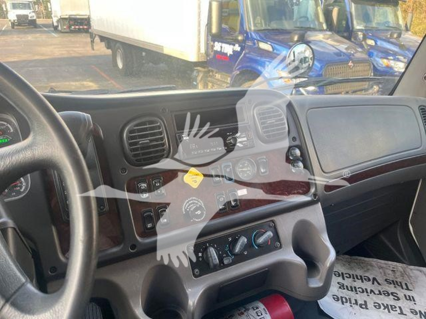 Vehicle dashboard interior of a Freightliner M2 106 cab showing steering wheel, HVAC controls, and instrument panel. Front windshield reveals moving van background.