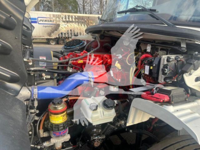 Freightliner M2 106 engine bay with visible components including red hoses, coolant reservoir, and wiring; multiple labels and caps readable.