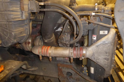 Truck engine bay showing intercooler hose, turbo, intake piping, and radiator support of a Freightliner Cascadia 113. Visible components include rubber hoses with clamps and metal piping.