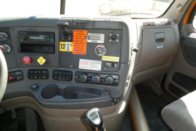 Dashboard of a Freightliner Cascadia cab showing control cluster, gauges, warning stickers, and gear shifter.