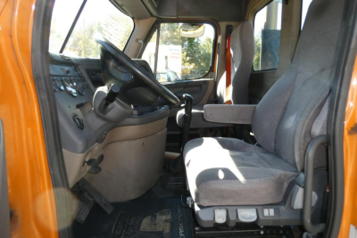 Dashboard and driver seat area of a Freightliner Cascadia 125 daycab showing steering wheel, gear shifter, and gray fabric seats.