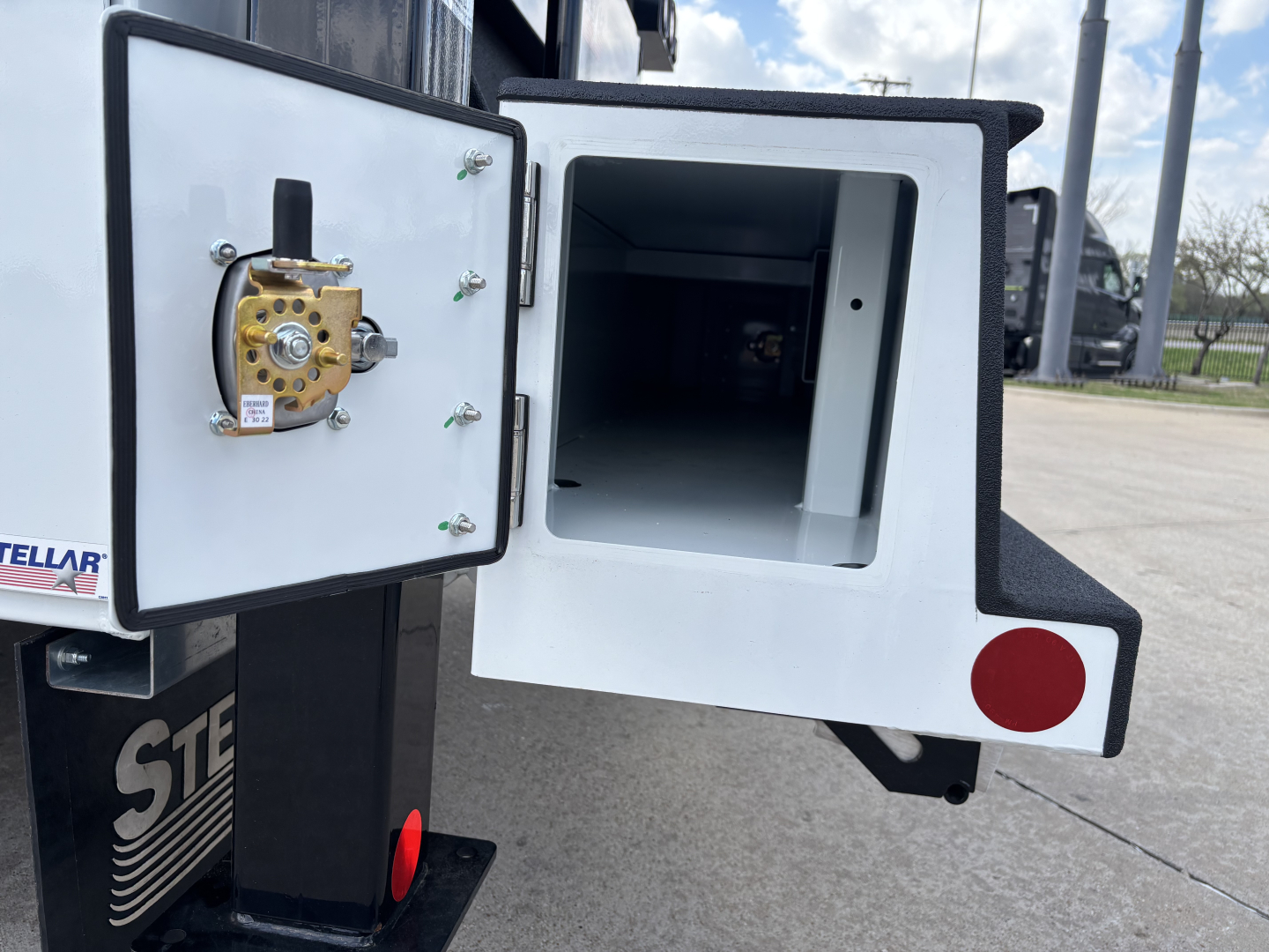 Rear of a 2025 Mack MD7 service truck showing a small insulated storage compartment with a door open, hinge hardware, and a round gold locking mechanism inside.
