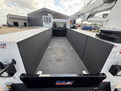 Truck bed of a 2025 Mack MD7 service/utility truck with open flatbed showing tall side panels and textured bed surface, Stellara branding on tailgate, surrounding equipment in background.