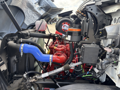Engine bay of new 2025 Mack MD7 showing red engine, blue radiator hose, black auxiliary tanks, and various hoses and wiring.