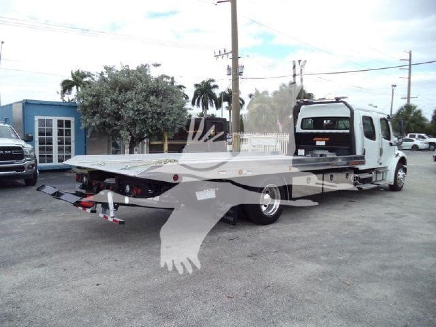 Freightliner M2 106 rollback truck in a dealership lot, long flatbed with tow winches and a ramp, cab in view, other vehicles and palm trees in background.
