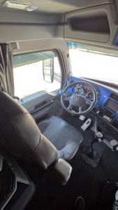Truck cabin interior showing steering wheel with Kenworth logo, dashboard with gauges, blue trim, gear shifter, driver seat, side window and overhead storage. No plates visible.