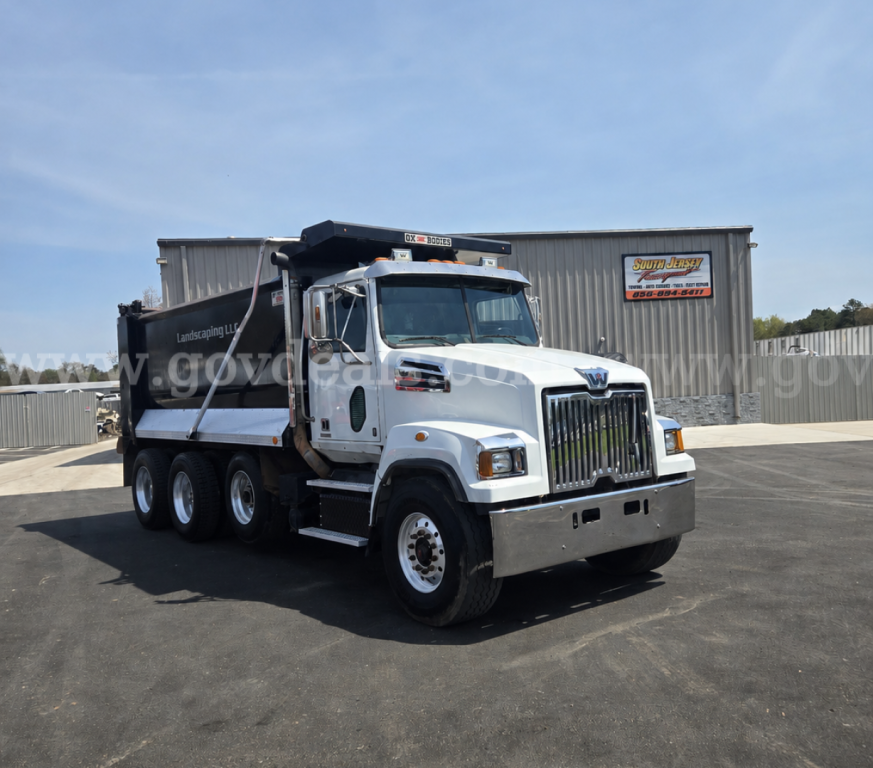 Truck showing Western Star 4700 dump body with dual rear axles and chrome grille. Visible text includes 'Lindstrom' on side of dump and a small sign on building behind.