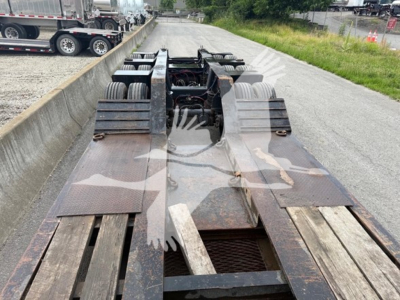 Lowboy trailer deck in view showing dual rear axles and wooden deck sections with metal framing and rusty surfaces.