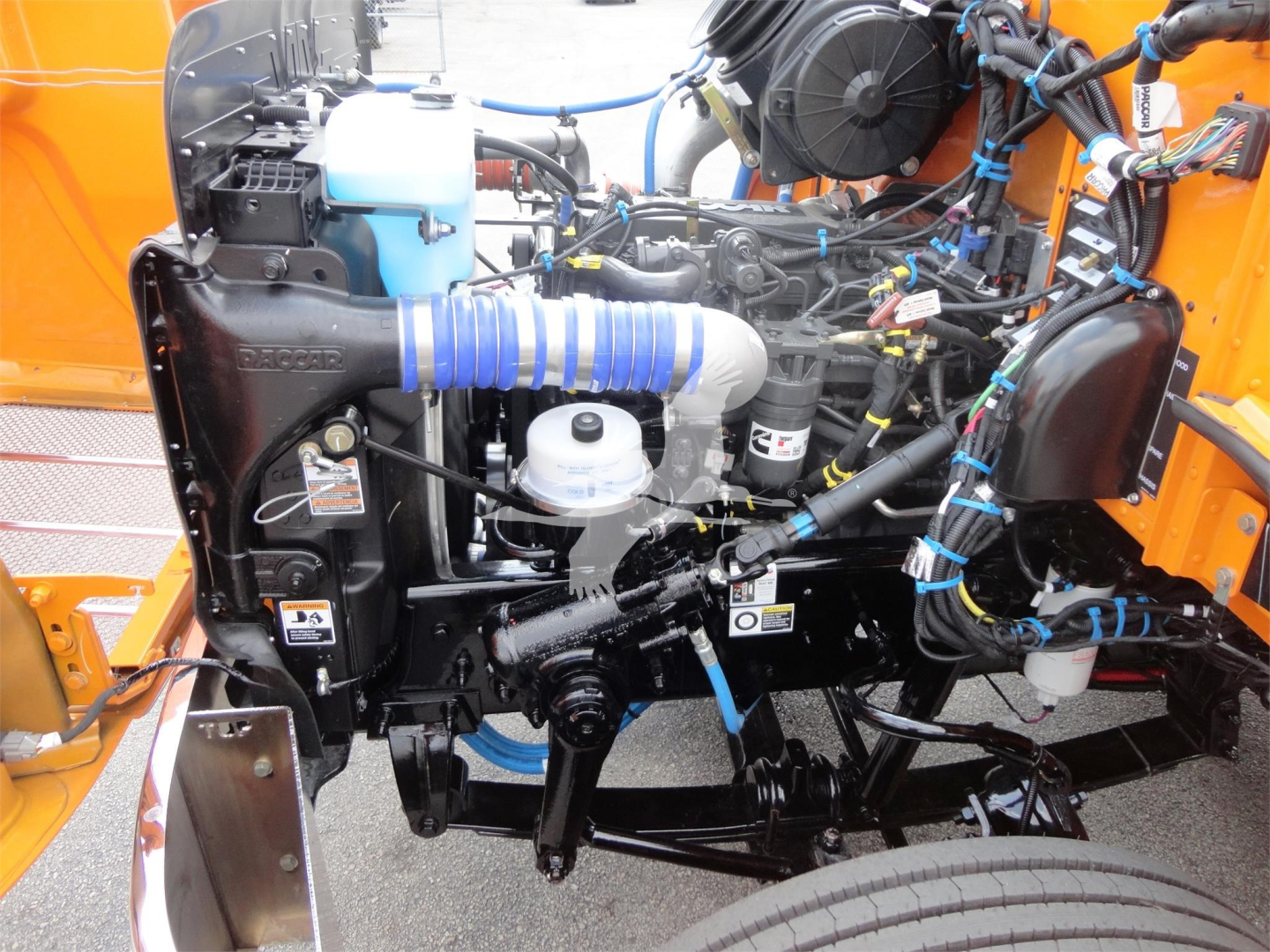 Peterbilt 337 engine bay with blue intake hose, coolant reservoir, and various hoses and wiring in an orange truck, showing engine components and front wheel.